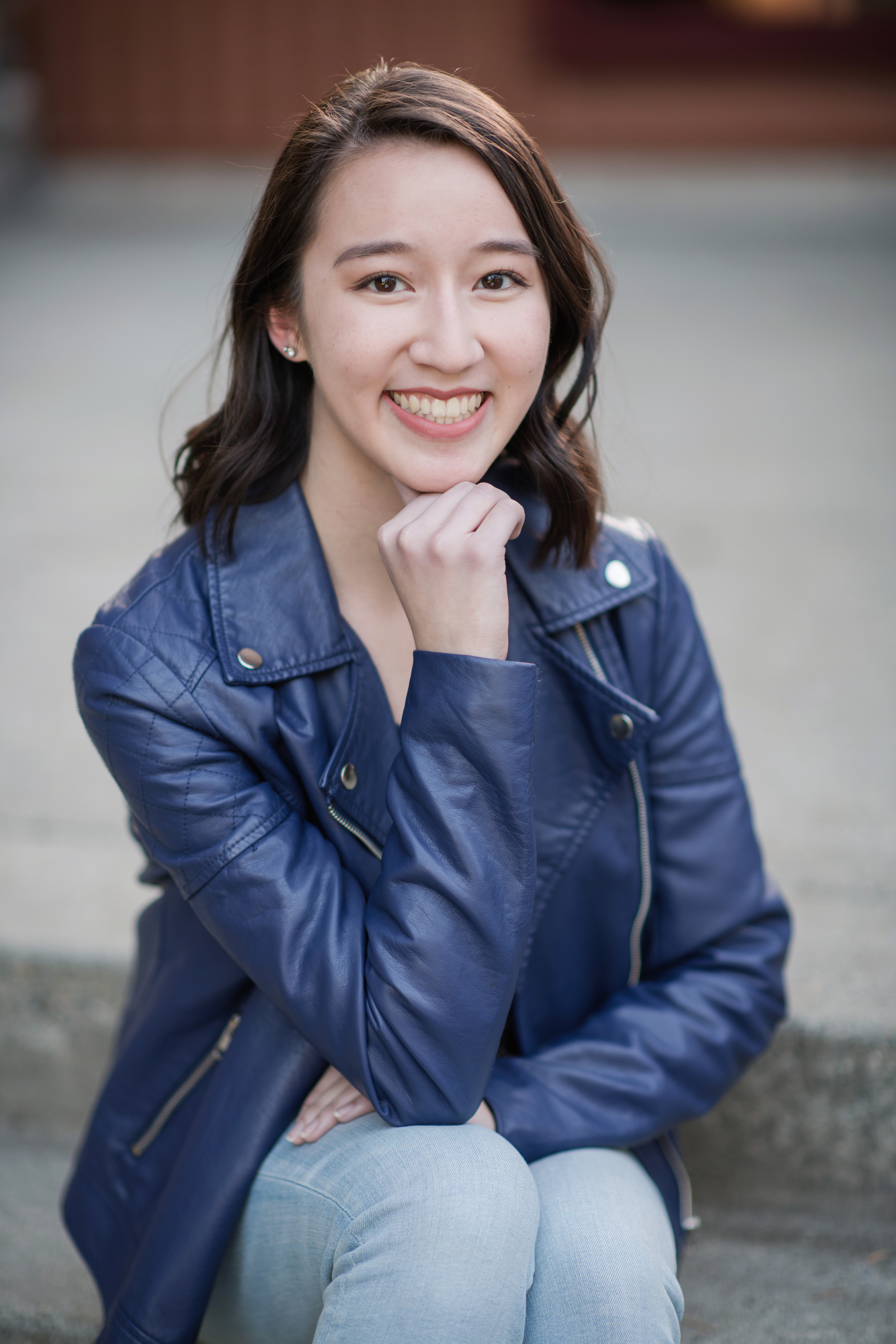 Portrait of Olivia smiling with her chin resting on her hand. She's wearing a blue jacket and is sitting on cement steps