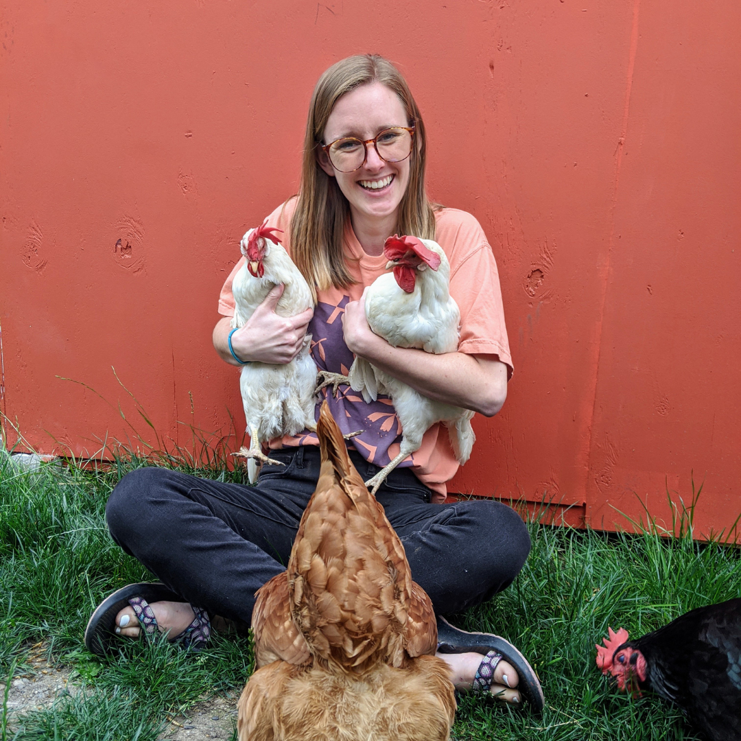 Hannah sits in the grass in front of a red wall, holding two chickens while others graze in the grass around her.