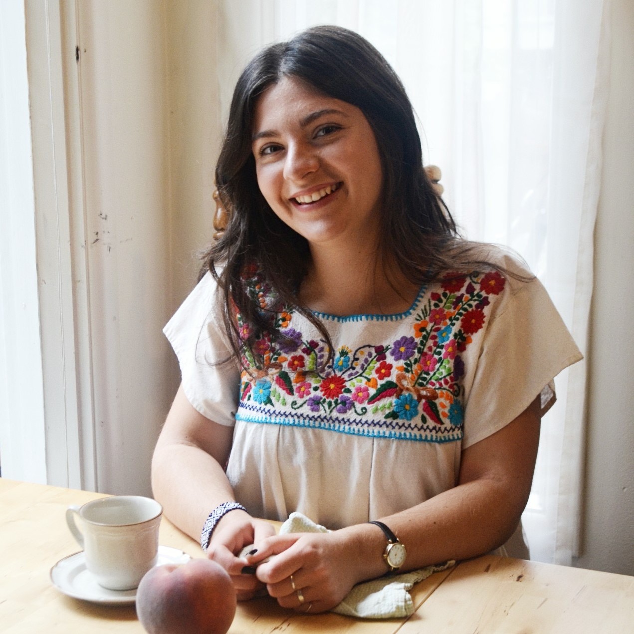 Serafima wearing an embroidered top sitting in soft natural light at a table with a coffee cup and a peach