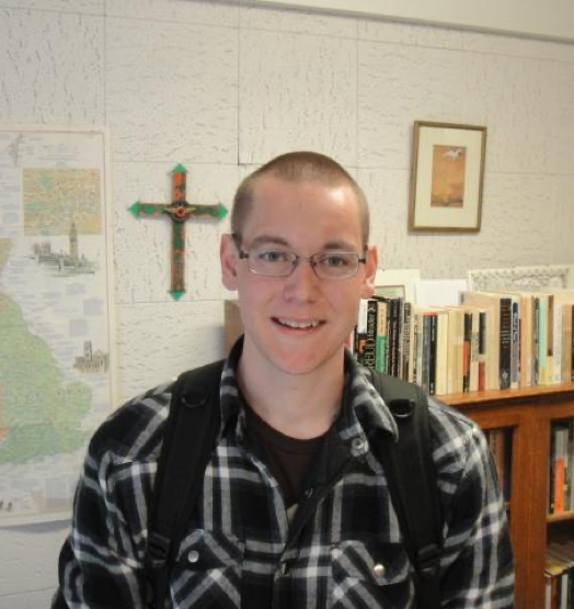 Portrait of Nathan in a flannel with a bookcase, map, and cross on the wall behind him