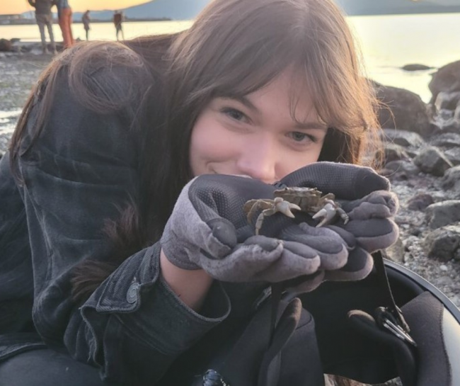 In gloved hands, Ula holds a small crab next to her face at the beach.