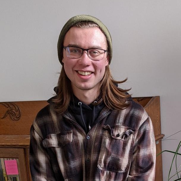 Portrait of Kai standing in front of a bookcase wearing a flannel and a beanie.