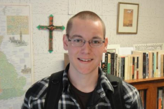 Portrait of Nathan in a flannel with a bookcase, map, and cross on the wall behind him