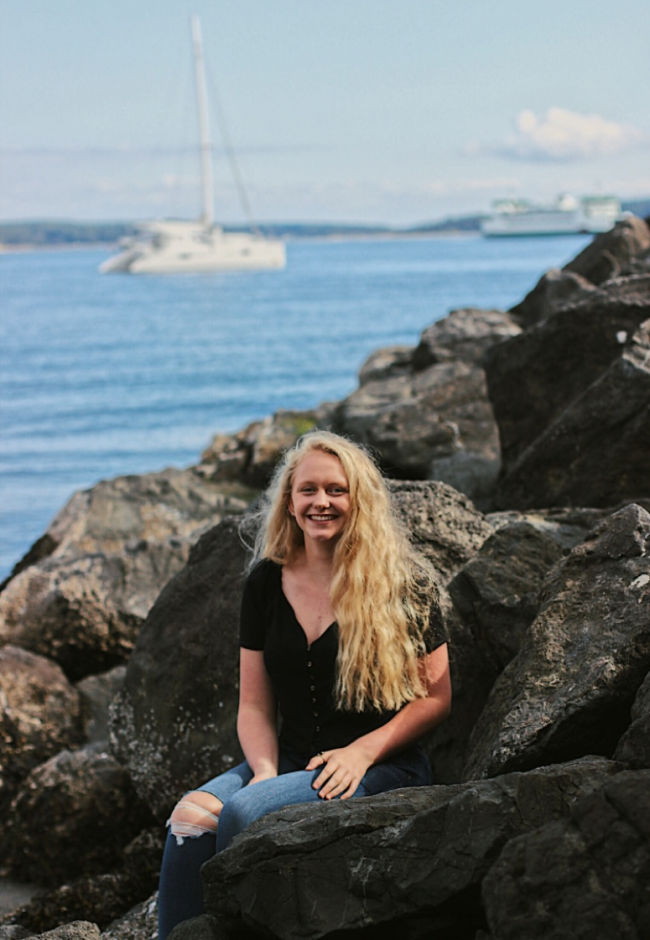 Photo of Allie sitting on rocks with a body of water and boats in the background