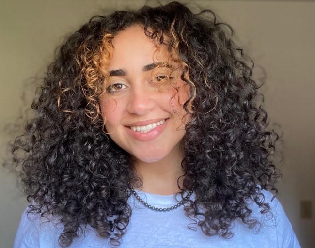 Portrait of Zoe smiling with her curly hair framing her face. She is wearing a blue shirt and a necklace.