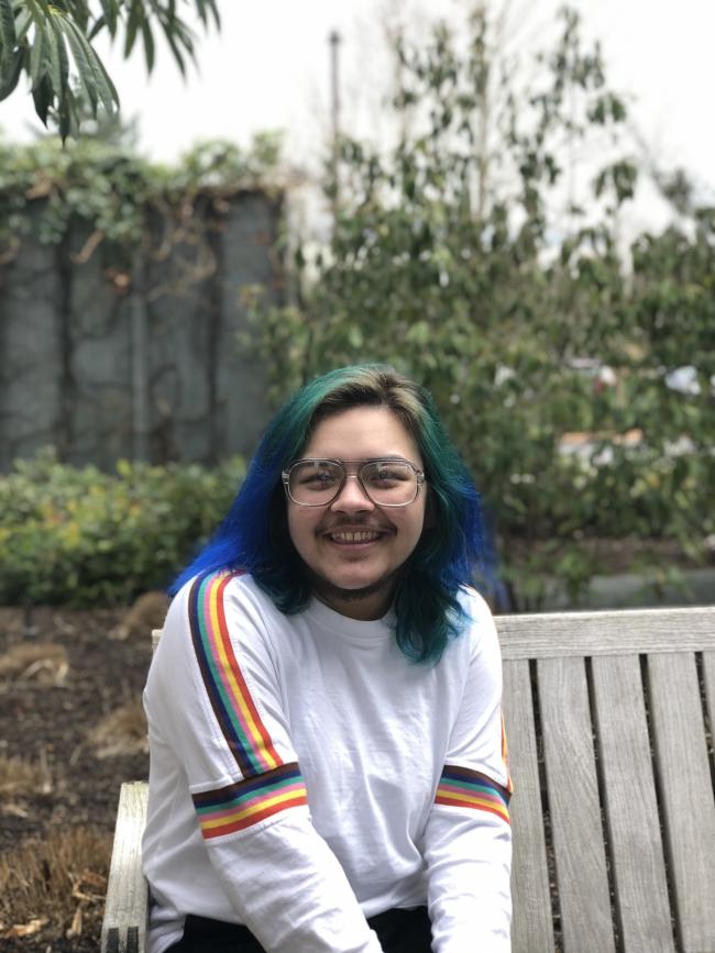 Portrait of Jay smiling and sitting on a bench. They are wearing a white long sleeved shirt with a rainbow stripe on the sleeves.